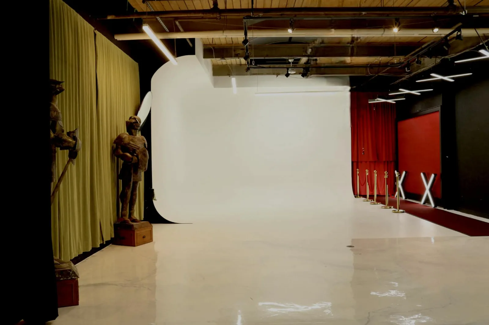 A wide, empty Toronto studio: a white cyclorama wall under a wooden ceiling with a lighting track, a gold curtain with two suits of armour standing against it on the left, and a red carpet behind brass stanchions along the right wall.